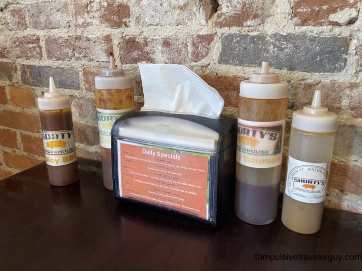 Shorty's Smokehouse sauce bottles including Spicy BBQ, Ginger, and Bourbon varieties flanking a napkin dispenser with the Daily Specials menu card against an exposed brick wall.