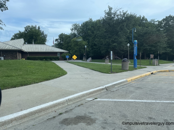 Highway rest area with a brick building, metal roof, Building Closed sign, blue emergency call box, walkways, and a small park with monuments visible behind.