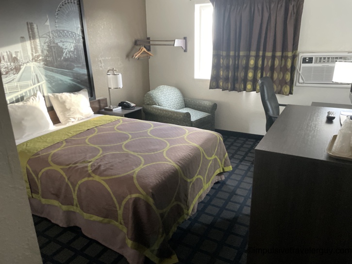 Super 8 motel room with a king bed covered in a brown-and-green circle-pattern bedspread, cityscape wall mural headboard, upholstered chair, desk, open closet rack, and a through-wall AC unit.