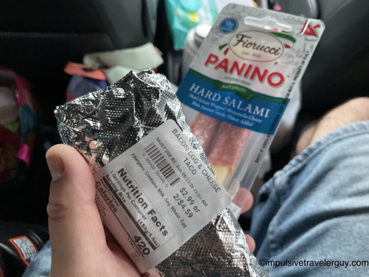 A hand holding a wrapped breakfast taco and a package of salami snacks in the back seat of the car.