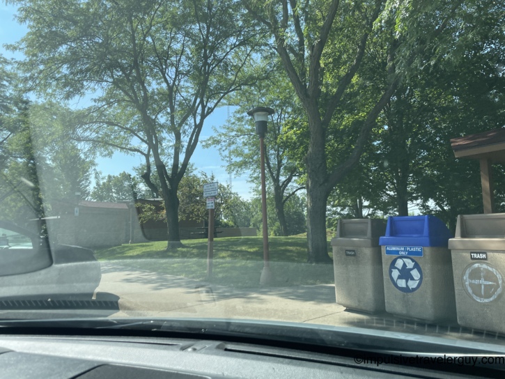 View from inside a vehicle looking out at a grassy US-151 rest stop with trees and outdoor waste bins.