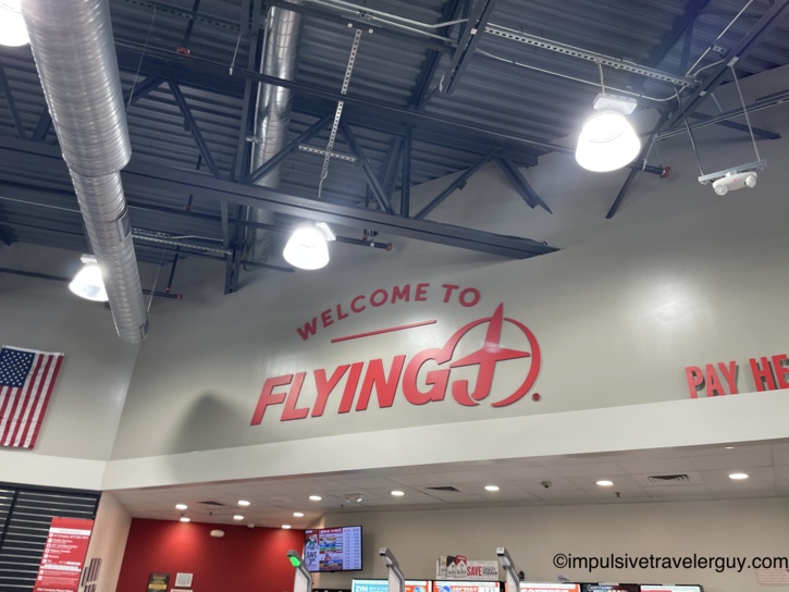 Interior of a travel plaza featuring a large red 'Welcome to Flying J' sign painted on the wall above the checkout counter.