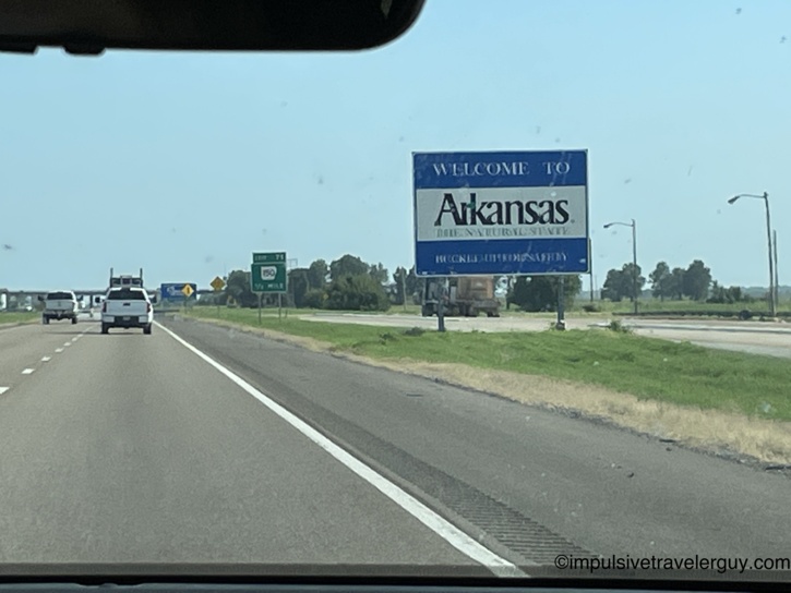 View through a car windshield driving on a highway, approaching a blue 'Welcome to Arkansas' state line sign.