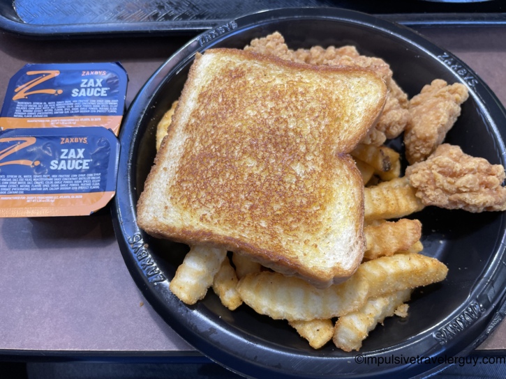 A Zaxby's meal in a black plastic bowl containing boneless chicken wings, crinkle-cut fries, and Texas toast, with packets of Zax Sauce on the side.
