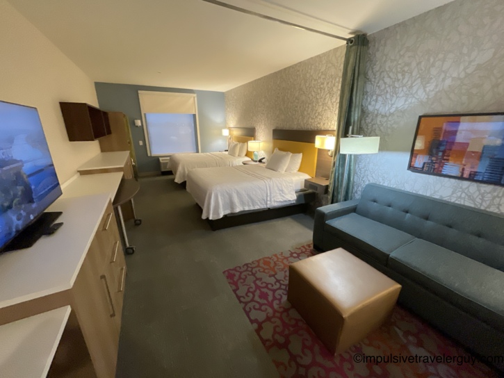 Interior of a North Little Rock Home2Suites hotel room featuring two beds with yellow headboards, a flat-screen TV, and a teal sofa.