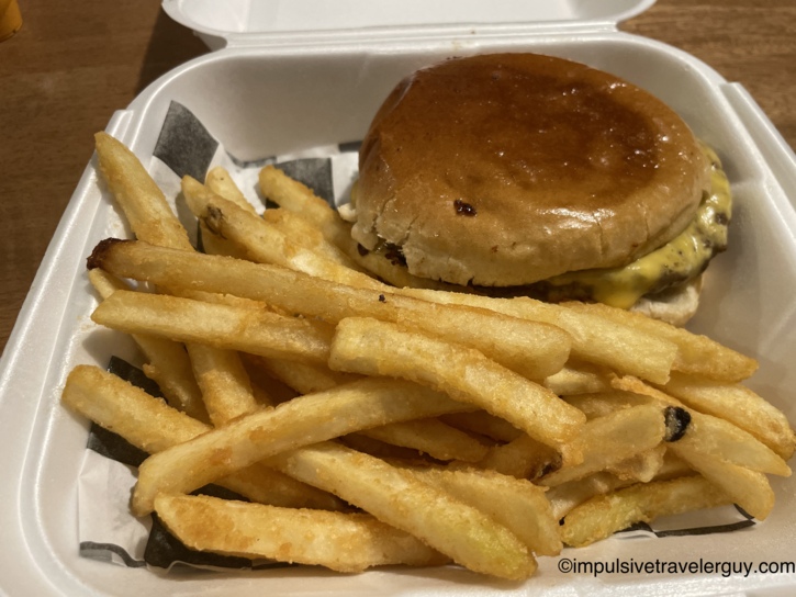 A cheeseburger with melted cheese and a side of french fries served in a white styrofoam takeout container from Taqueria Los Olivos.