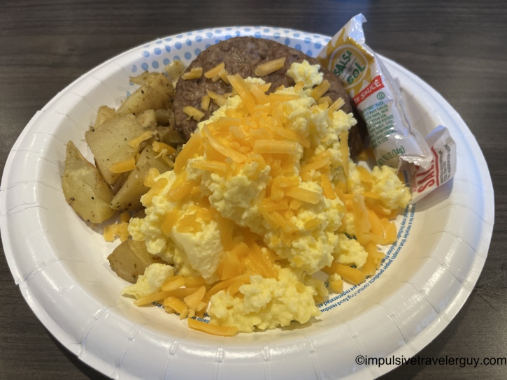 A hotel breakfast plate filled with scrambled eggs topped with shredded cheddar cheese, seasoned diced potatoes, a sausage patty, and packets of salsa.