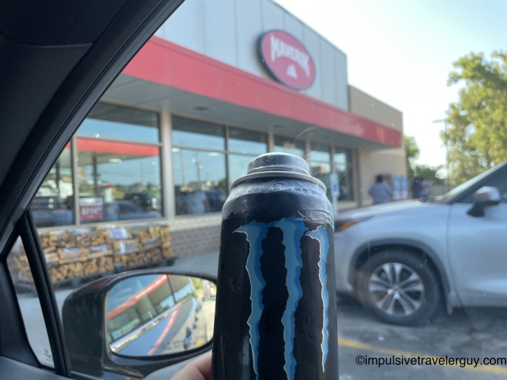 A hand holding a cold Monster Energy drink inside a car, with a Maverik gas station storefront visible through the window.