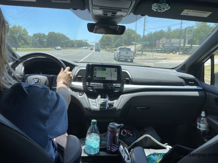 View from the back seat of a car showing a woman driving into a sunny Texas town, with water bottles resting in the center console area.