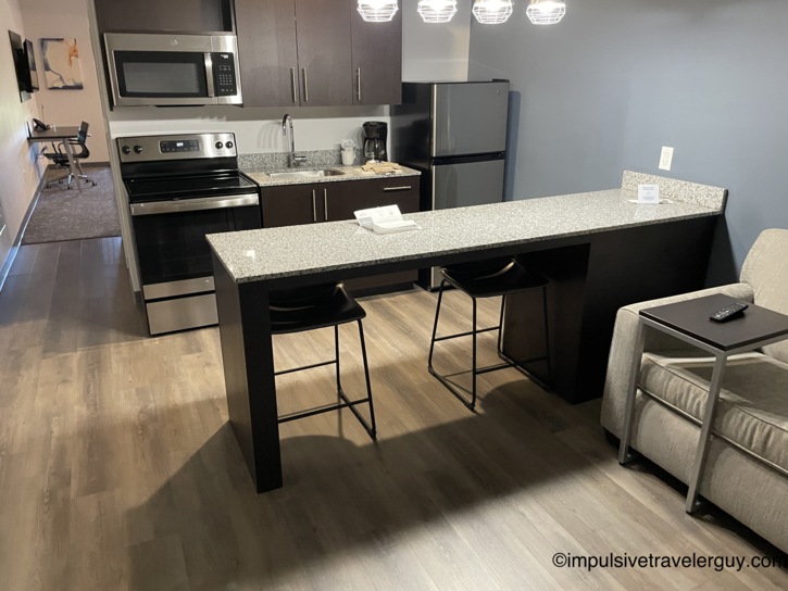 Interior of a hotel suite kitchen featuring full-size appliances, a granite peninsula with bar stools, and an open living area.
