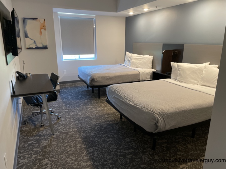 A separate hotel bedroom containing two beds, a dark wood desk, and a window with a drawn shade.