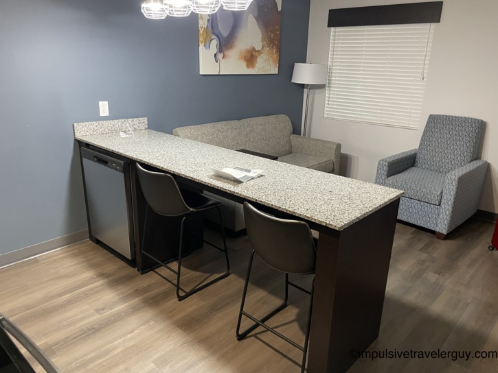 A hotel suite living area featuring a grey sofa and a patterned armchair next to the granite kitchen peninsula.