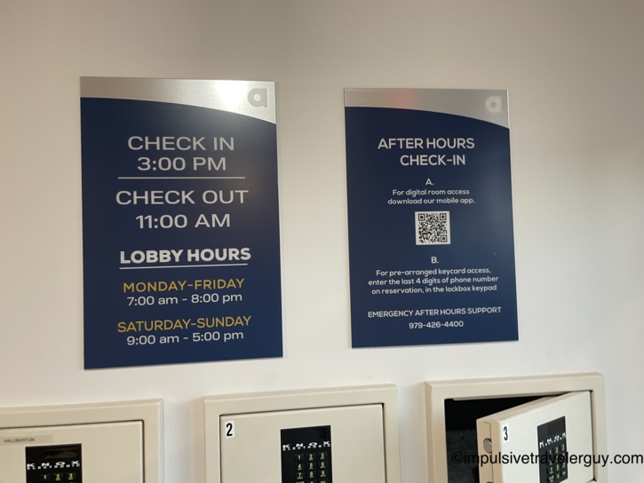 Hotel wall signs detailing limited lobby hours and after-hours check-in instructions above a row of electronic key lockboxes.