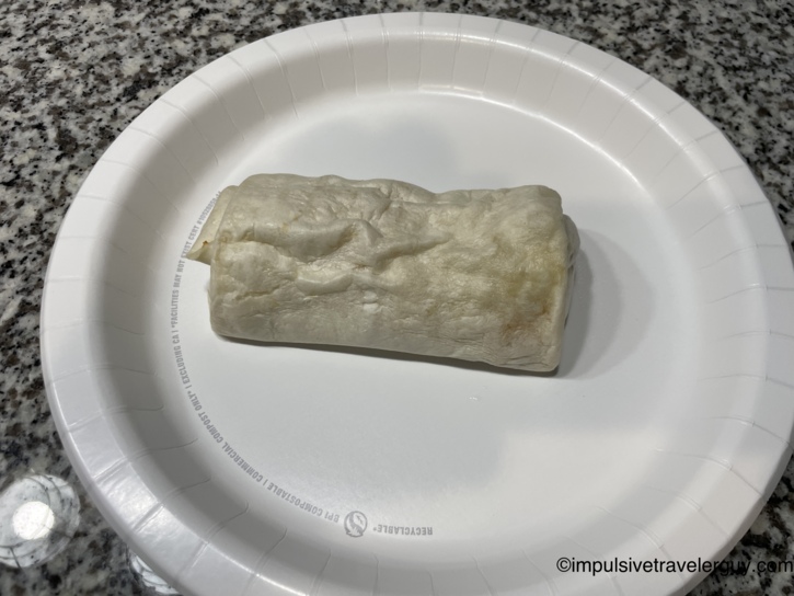 A plain wrapped breakfast burrito resting on a white paper plate on a granite countertop.