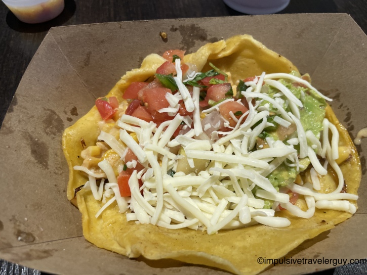 A taco from Fuego served in a cardboard food boat, layered with corn, diced tomatoes, guacamole, and shredded white cheese.