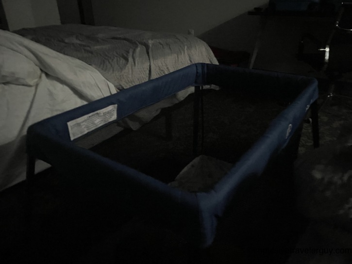 A dimly lit hotel room showing a blue Pack 'n Play travel crib set up for nap time beside a large bed.