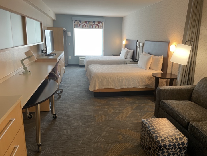 Spacious interior of an Austin Home2Suites hotel room featuring two beds, a long wooden desk unit, and a dark grey sofa.