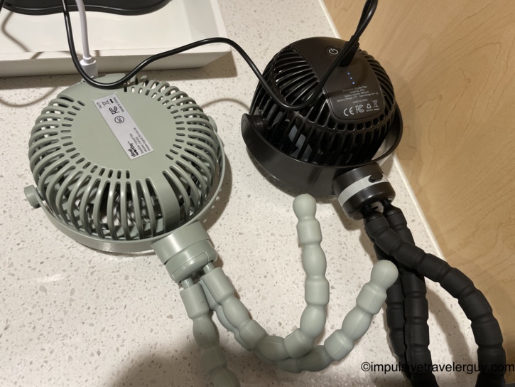Two small battery-powered portable fans charging via USB cables on a hotel room countertop.