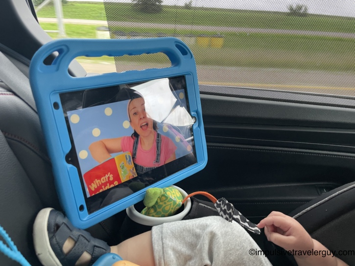 An iPad in a blue protective case mounted to a car headrest playing a Ms. Rachel video to entertain a child sitting in a car seat.
