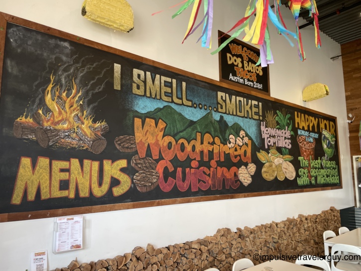 A large, colorful chalkboard mural inside Dos Batos Tacos reading 'I Smell... Smoke!' positioned above a decorative stack of chopped firewood.