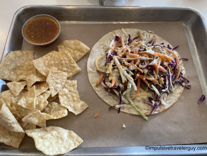 A metal tray holding a Dos Batos shrimp taco topped with slaw and sauce, served next to tortilla chips and salsa.