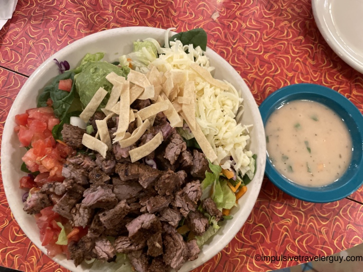 A Chuy's taco salad bowl filled with grilled steak, lettuce, tomatoes, guacamole, and tortilla strips, served with a side of creamy dressing.