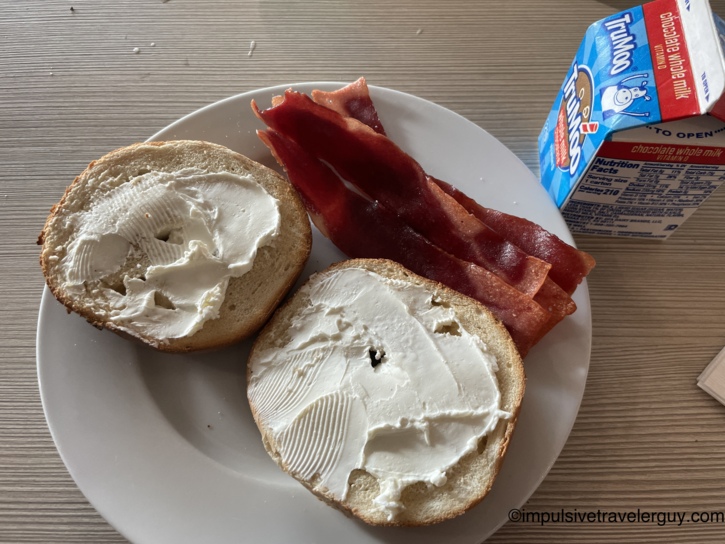 A lighter breakfast plate holding a bagel spread with cream cheese and strips of bacon, accompanied by a carton of chocolate milk.