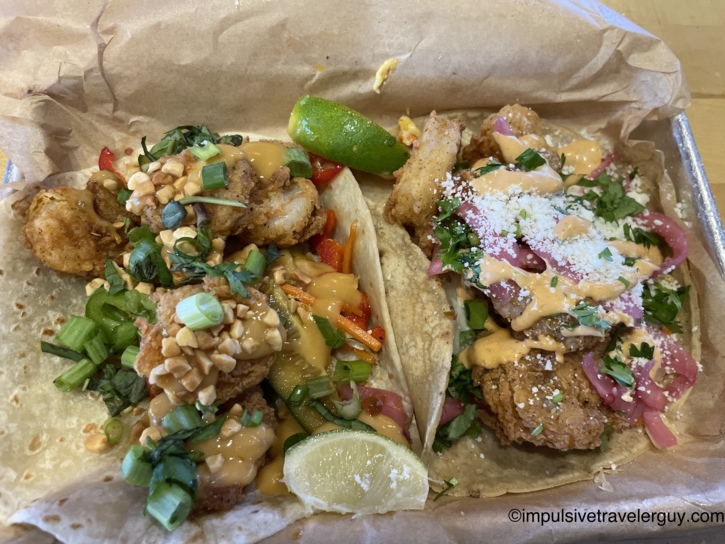 Two gourmet Torchy's Tacos holding fried shrimp, fresh garnishes, and creamy sauce served on brown paper in a metal tray.