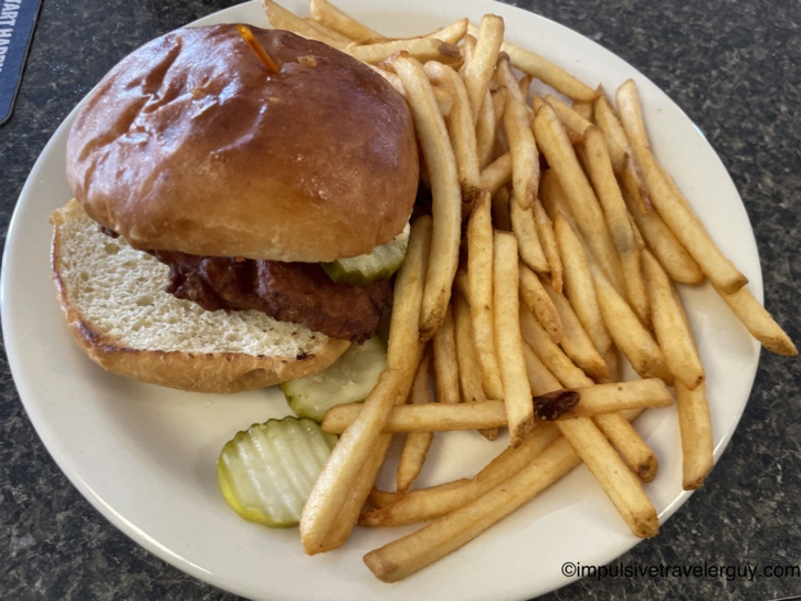 A fried chicken sandwich with pickles on a toasted bun, served alongside a portion of french fries on a round white plate.