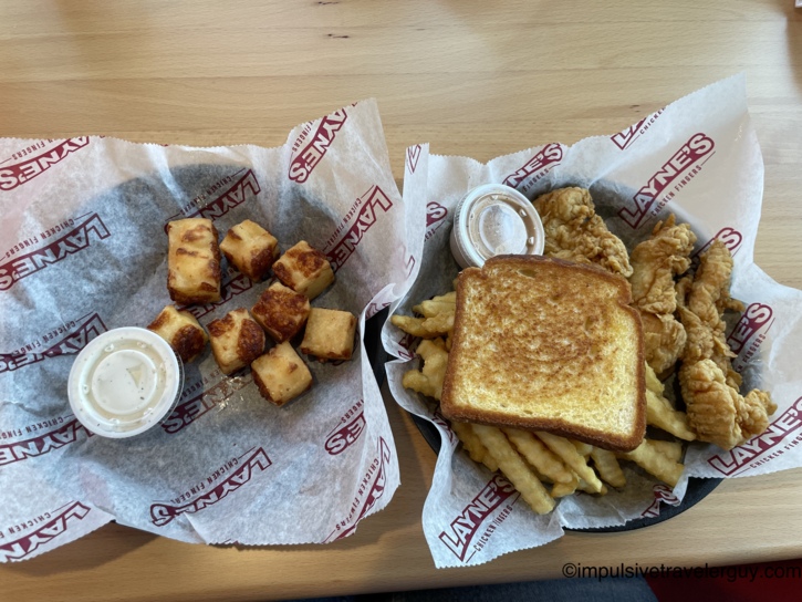 Two baskets of food resting on Layne's branded paper. The basket on the left contains square fried cheese nuggets and a container of dipping sauce. The basket on the right contains fried chicken fingers, crinkle-cut french fries, a piece of Texas toast, and dipping sauce.
